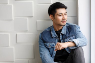 Cilveli kot ceketli Asyalı genç adam beyaz duvara karşı dost canlısı ve gülümseyen hareketlerle oturuyor. Moda şehir portresi konsepti