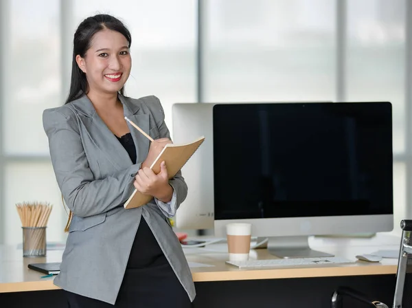 Young attractive Asian woman in grey business suit writing on notebook in modern looking office with blurry windows background. Concept for modern office lifestyle.