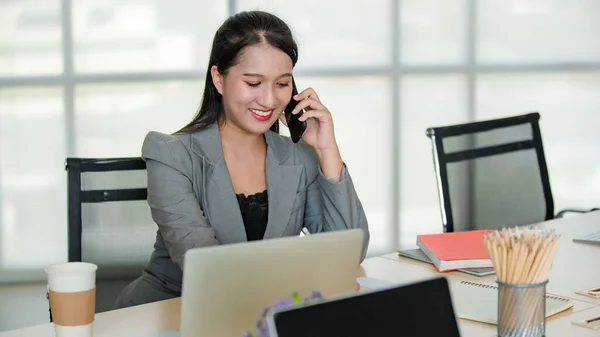 Young attractive Asian woman in grey business suit sitting talking on mobile phone in modern looking office with blurry windows background. Concept for modern office lifestyle.