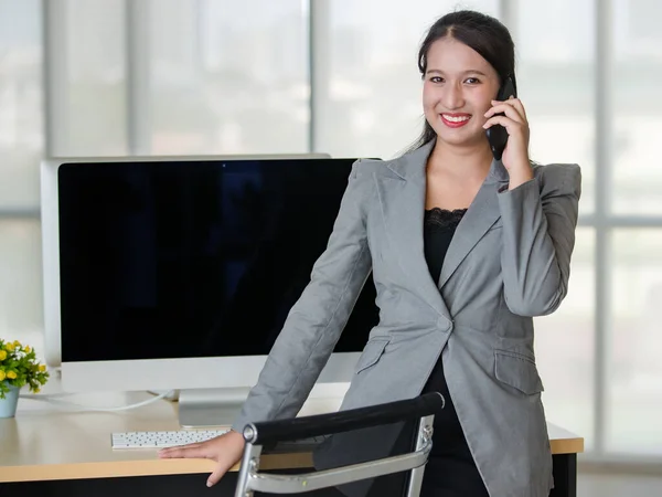 Young attractive Asian woman in grey business suit talking on mobile phone in modern looking office with blurry windows background. Concept for modern office lifestyle.