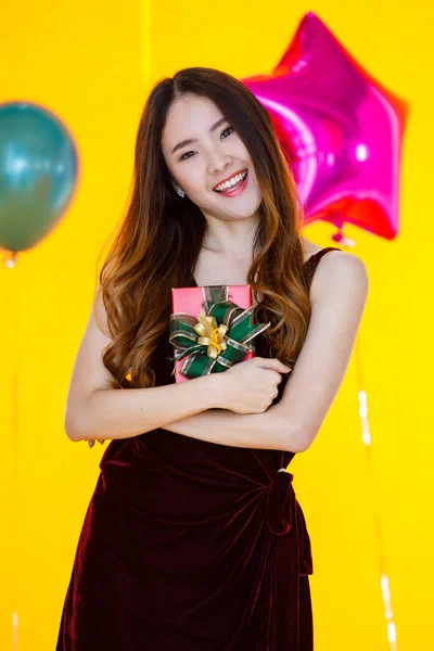 アジアの美しいです女の子でセクシーなドレス立って笑顔グリーンリボンプレゼント赤いボックスで手で前カラフルな風船で黄色の背景に驚き誕生日パーティー — ストック写真