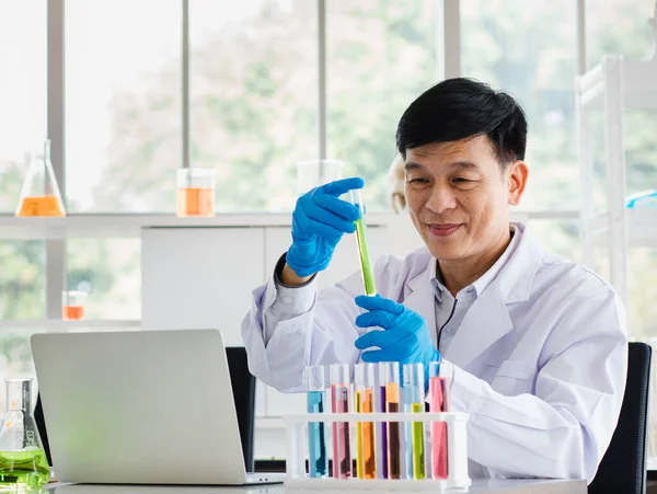 2 asian men are scientist holding and look at test tube with chemical liquid. Focus to young scientist. Microscope, laptop and more test tubes with liquid put on table in laboratory room in the morning.