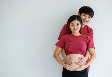 Gülen genç karı ve koca beyaz arka planda yan yana duruyorlar. Bir erkek hamile bir kadını kucaklar, elini karnına koyar. İnsanlar, aile ve yaşam tarzı konsepti. Boşluğu kopyala.