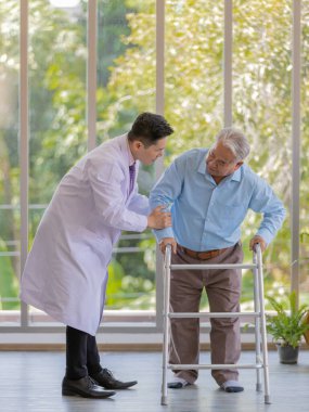 Beyaz laboratuvar önlüğü giyen genç bir Asyalı erkek doktor sağ kolunu tutarak hastasına destek olmaya çalışırken açık mavi gömlek ve kahverengi pantolon giyen yaşlı, şişman, gri saçlı bir hasta ayakta durarak hastaneye yürümek için Walker 'ı kullanmaya çalışıyor..
