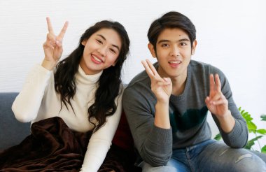 Beyaz ve gri uzun kollu sweatshirt ve kotlu şirin gülümseyen Asyalı sevgili portresi kanepeye oturur, kameraya bakar ve zafer işaretiyle parmaklarını gösterir..