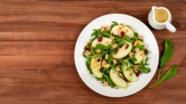 Roketli yeşil elma dilimi salatası, yulaf tanecikleri, kurutulmuş kızılcık ve ağaçlık masada beyaz kabın içine yerleştirilmiş kaju fıstığı. Yağ sirkesi salatası sosu, meze. Dekorasyon olarak kullanılan iki roket yaprağı.