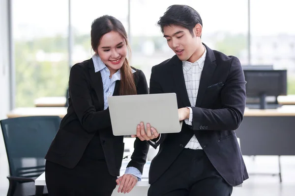 Happy young businessman holding laptop and pointing at the computer screen talking with his lovely female colleague in the workplace, exciting on a business achievement