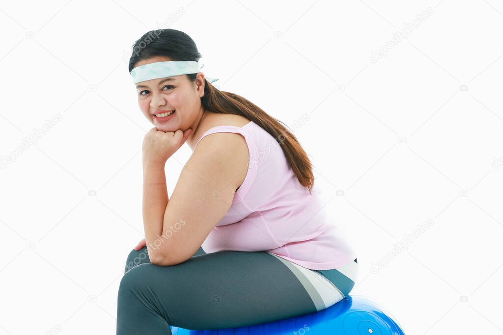 Portrait isolated studio shot of Asian happy healthy strong big fat girl in sport clothing leggings with headband sit smiling on blue exercise fitball rest chin on hand in front white background.