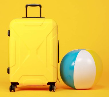 Siyah saplı sarı valiz ve sarı arka planda mavi ve sarı plaj topu. Plaj, seyahat ve tatil kavramı.