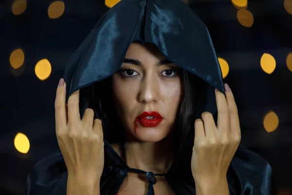 Young Asian vampire female in Halloween wear with black hood on head and red lips with dripping blood looking at camera against dark background with blurred lights