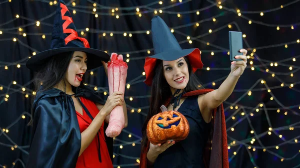 万圣节庆祝活动期间 戴着女巫帽 头戴南瓜头的时髦少女们一边在智能手机上自拍一边玩 — 图库照片