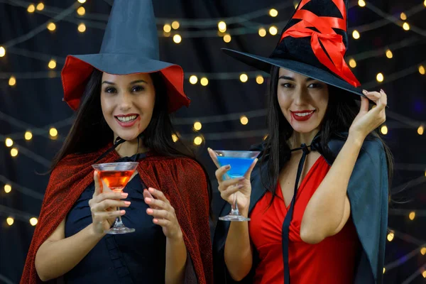 万圣节庆祝活动期间 穿着巫婆服装和帽子的时尚年轻女性朋友一边喝着五颜六色的酒精饮料 一边开心地微笑着 — 图库照片