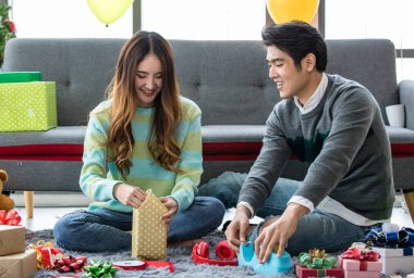 Romantik genç Asyalı çiftler oturma odasındaki kanepenin yanında oturup gülümseme ve kahkahalarla güzel hediye paketleri hazırlıyorlar.