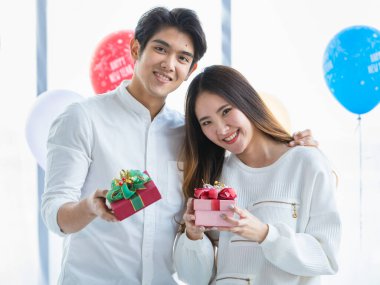 Beyaz tişörtlü romantik genç Asyalı çiftler birlikte mutlu mesut duruyorlar, gülümsüyorlar ve güzel hediye kutlarını yeni yıl partisinde süslü balonlarla süslenmiş tatlı aşkı kutlarken gösteriyorlar.