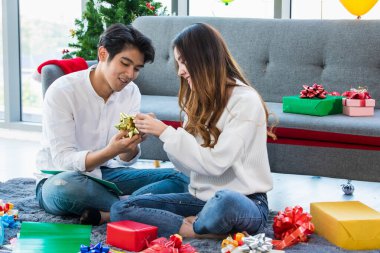 Romantik genç Asyalı çiftler, mutlu bir gülümseme, oturma odasında birlikte oturup, kış festivalinde Noel partisi için hediye paketi hazırlamak için birbirlerine kurdele hazırlamak için yardım etmek.