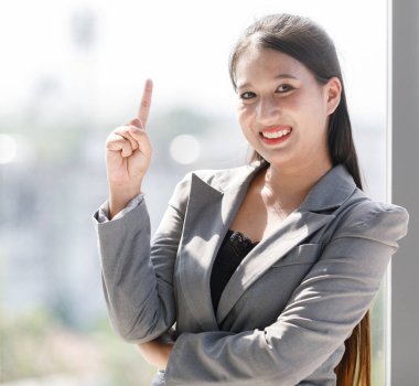 Gri takım elbiseli Asyalı gülümseyen kadın ofis penceresinde dikiliyor. İş kadını tam boy ayakta duruyor. Güzel ve kendine güvenen çalışan bir kadın.