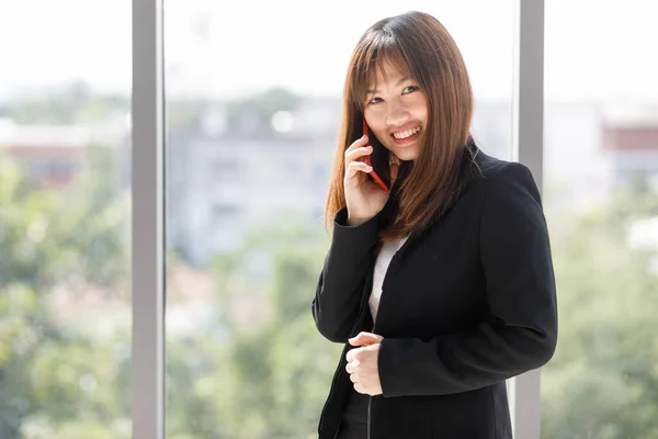 亚洲笑容满面的女人手握智能手机 一身黑色西服 女商人在办公室通过电话谈论工作 美丽自信的职业女性 — 图库照片