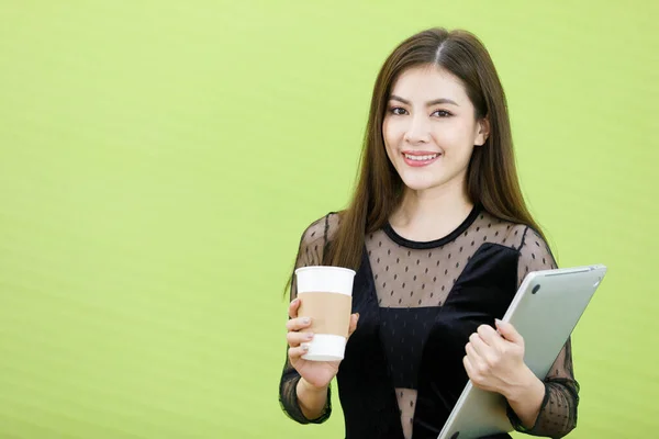 Young and beautiful Asian female secretary holding coffee cup and paper notebook on green background with smile face.