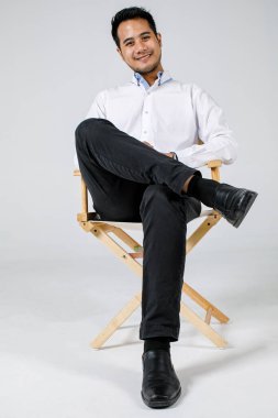 Genç Asyalı iş adamı stüdyo koltuğunda oturuyor kendinden emin ve mutlu, beyaz arka planda izole edilmiş bir portre..