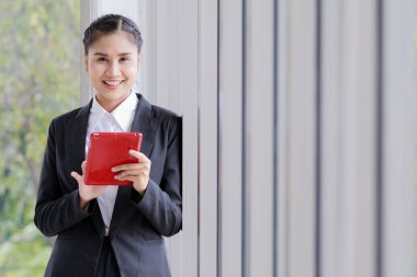 Asyalı başarılı iş kadını resmi takım elbiseli kadın girişimci duvara yaslanmış gülümse kameraya bak dokunmatik ekran kırmızı tabletine bak müşteri e-postalarını kontrol et.