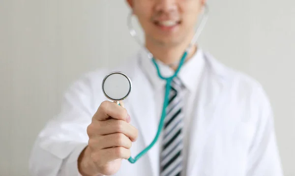 Closeup shot of stethoscope in hand of male doctor wears white lab coat uniform stand smiling in blurred background reach out to listen and examine patient heartbeat in ward monitor room of hospital.