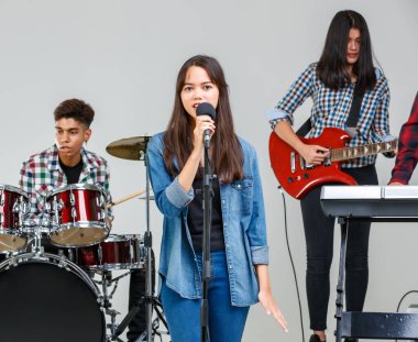 Müzik çalan ve birlikte şarkı söyleyen gençlerin grup fotoğrafı. Genç öğrenciler elektro gitar, davul ve klavye çalarlar. Arka planda bir müzisyenle şarkı söyleyen genç bir bayan şarkıcı.