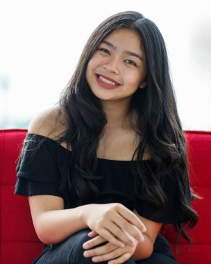 Siyah elbiseli, güzel, gülümseyen genç bir kızın, kanepede oturup stüdyodaki kameraya bakarken ön manzara portresi. Balo partisindeki güzel ergen kız.