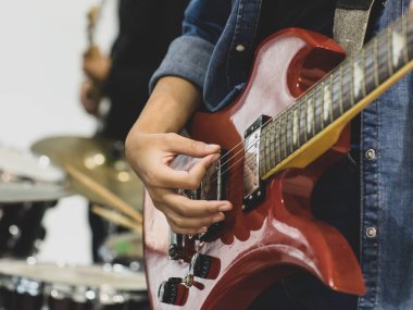 Genç bayan müzisyen ellerinin elektro gitar çalarkenki yakın çekimi. Arka planda davul olan gitaristin ellerine odaklan. Gitarist sınıf arkadaşıyla birlikte müzik çalıyor.