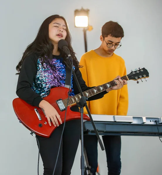 Portrait shot of a young teenage singer singing a song with a microphone and playing the electric guitar. Professional junior student vocalist practicing with male pianist in the background.
