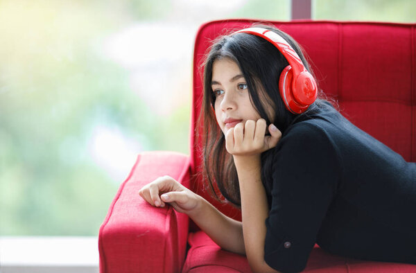 Portrait shot of cute smiling young Thai-Turkish teenager enjoying listening to music with headset. Young mixed-race junior girl model lying down on the red couch with hand to chin gesture