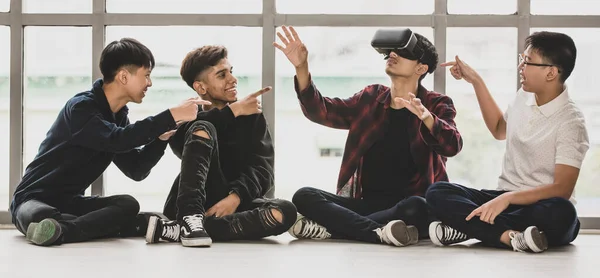 Group of four handsome young male teenagers with cute smiling sitting on floor together. Junior boy playing virtual reality game with a headset with friends pointing to him. Concept of technology