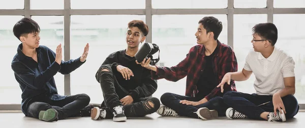 Group of four handsome young male teenagers with cute smiling sitting on the floor together. Junior boy raising hands up to deny playing virtual reality headset that was given by his friends