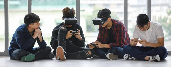 Group of four handsome young male teenagers with cute smiling sitting on floor together. Junior boys playing games with virtual reality headset, controllers, and smartphones. Concept of technology