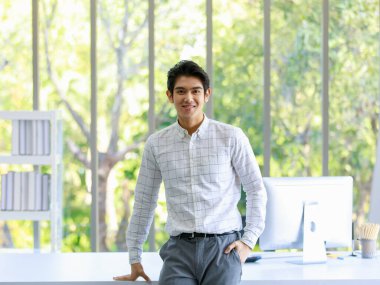 Asyalı başarılı beyaz yakalı erkek memurun, çalışma masasına yaslanıp bilgisayar monitörüyle gülümseyen kameraya bakıp elini pantolonunun cebine soktuğu bir portre resmi..