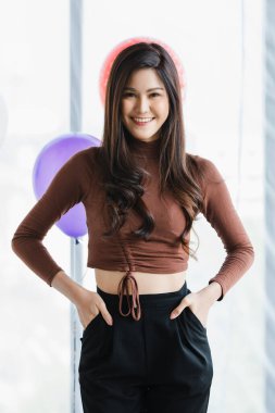 Asyalı, mutlu, uzun kollu, kahverengi tişörtlü, gülümseyen bir kadının portre çekimi. Kameraya bak. Öndeki renkli balonların ceplerine ellerini koy..