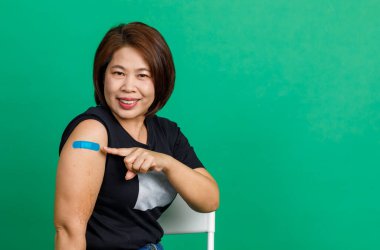 Asyalı orta yaşlı kadının stüdyo fotoğrafı. Gülümse ve kolunda mavi alçı bandajı göster. Coronavirus covid 19 aşı aldıktan sonra yeşil arka plandaki klinikteki doktor tarafından..