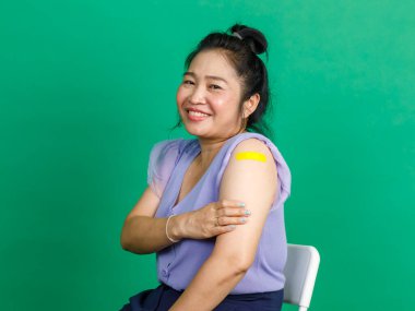 Stüdyoda çekilen fotoğrafta, mor şifon elbiseli, sarı bandaj bandını gösteren kameraya gülümseyen, orta yaşlı, mutlu, topuz saçlı kadın yeşil arka planda Coronavirus covid 19 aşısı yaptırıyor..