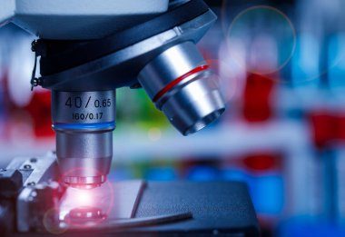 Bilimsel mikroskop büyütme merceğinin yakın plan görüntüsü cam plaka numunesinde hastane laboratuarındaki bulanık ve mavi test tüpleri önünde kırmızı ışık parlaması ile görüntüleniyor..