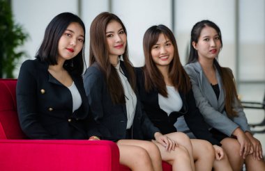 Resmi takım elbiseli dört çekici Asyalı bayan ofis arkadaşı ofiste kırmızı koltukta oturmuş kameraya gülümsüyor. Modern ofis çalışması kavramı.