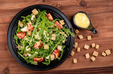 Domatesli roket salatası, kızarmış tavuk, siyah tabakta parmesan peyniri, çuval bezinin üzerinde mayonez dolu bir sos kabı ve ahşap masadaki ekstra kızarmış ekmek..