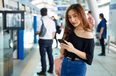 Gülümseyen sevimli ve aktif Asyalı bayan yolcu metroda akıllı telefonlu bir şekilde kameraya bakıyor. Modern şehir yaşamının seyahat ve iletişim teknolojisi fikri