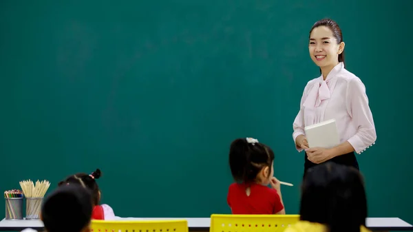 アジアの若い美少女先生のポートレート撮影教室で小学生に授業をしながら 手で教科書を持ってカメラを見て立って講師 — ストック写真