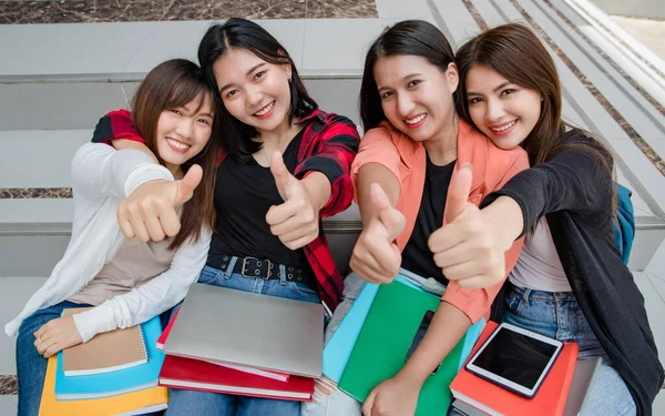 Group of four young attractive asian girls college students smiling to camera doing thumbs up in university campus outdoor. Concept for education, friendship and college students life.