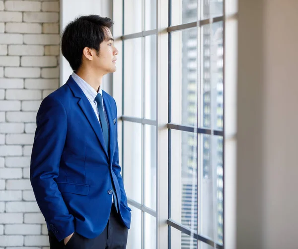 Portrait closeup shot of Asian young intelligence college teen student wears blue formal suit uniform and necktie standing hold hands in pants pockets look outside glass windows in front brick wall.