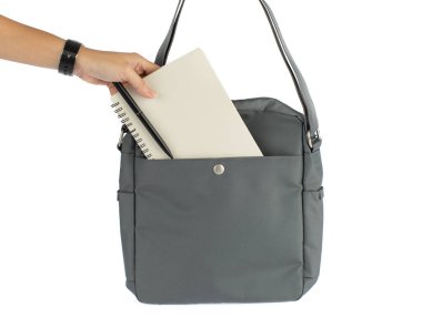 Kadın elinin izole edilmiş yakın çekim görüntüleri defter ve kalemi beyaz arka planda uzun omuz askısı olan küçük şehir iş kadını gri çantasının ön cebine koyuyor..