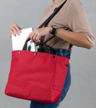 Closeup studio shot of female model in brown long sleeve shirt putting silver notebook laptop computer into compartment pocket of trendy urban big red crossbody unisex handbag on gray background. clipart