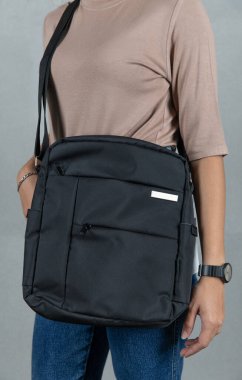 Kahverengi uzun kollu gömlekli ve kot pantolonlu kadın modelin yakın çekim stüdyo resmi. Gri arka planda uzun omuz bandı olan modaya uygun, siyahi, çok amaçlı uniseks çantası asılı duruyor..