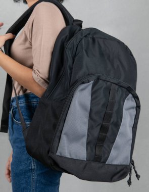 Kahverengi uzun kollu gömlekli ve kotlu kadın modelin yakın plan stüdyo fotoğrafı. Moda olan şehir siyah ve gri renkli uniseks seyahat sırt çantasını gri arka planda asılı duruyor..