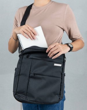 Kahverengi uzun kollu tişörtlü kadın modelin yakın çekim stüdyosu gümüş dizüstü bilgisayarı gri arka planda büyük siyah çapraz gövdeli uniseks çantasının cebine koyuyor..