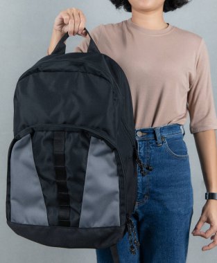 Kahverengi uzun kollu gömlekli ve kot pantolonlu kadın modelin yakın çekim stüdyo resmi. Gri arka planı önünde uzun omuz askısı olan, modaya uygun, büyük siyah, uniseks çantasının sapını tutuyor..